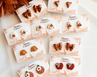 Könnte beinhalten: Eine Sammlung handgefertigter Polymer Clay Ohrringe. Die Ohrringe zeigen verschiedene herbstliche Designs, darunter Schleifen, Kürbisse, Pullover und Äpfel. Jedes Paar ist auf einer Karte mit dem Text "happy Cutter Co." präsentiert.