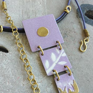 May include: A handcrafted necklace featuring a lavender pendant with gold accents. The pendant is composed of three connected rectangular pieces with a leaf pattern and a gold-colored chain. The necklace has a lavender cord.