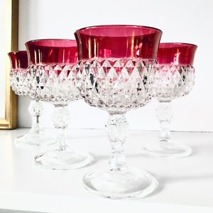 Peut inclure: Quatre verres à vin en cristal transparent avec des bords rouge rubis. Les verres présentent un motif taillé en diamant sur le bol et reposent sur un pied et une base transparents. Les verres sont disposés sur une surface blanche.