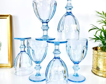 May include: A collection of five light blue glass goblets with vertical ribbed detailing on the bowls and stems. Some glasses are standing upright while others are inverted, showcasing the circular bases and the consistent pale blue hue.