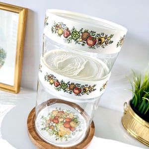 May include: Clear glass canisters with white lids and a wooden base. The lids and base feature a colorful fruit and vegetable design. The canisters are stacked, showcasing the decorative pattern and the clear glass construction.