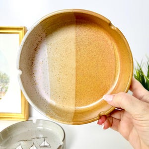 May include: A round ceramic ashtray with a two-tone design. One half is a speckled off-white, and the other is a warm, golden-brown. The ashtray has a curved rim with a notch for resting cigarettes. Another ashtray with a sailboat design is visible.