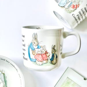 May include: White ceramic mug with illustrations of rabbits, reminiscent of a children's story. The mug has a curved handle and text. Other items with similar illustrations are also visible.