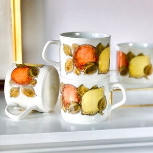 May include: Three white ceramic mugs with a fruit design. The mugs feature a pattern of oranges and lemons with green leaves. The mugs are stacked on top of each other.
