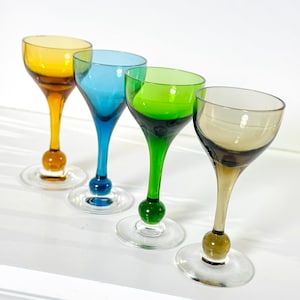 May include: Four colorful stemmed glasses in yellow, blue, green, and brown. Each glass has a unique color, a rounded base, and a clear, circular foot. The glasses are arranged in a row, showcasing their vibrant hues and elegant design.