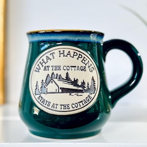 May include: A dark green ceramic mug with a glossy finish and a blue rim. A circular cream-coloured emblem on the front features a black line drawing of a cottage surrounded by trees and the text "WHAT HAPPENS AT THE COTTAGE STAYS AT THE COTTAGE."