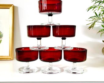 May include: A set of six ruby red glass dessert bowls arranged in a pyramid stack. Each bowl features a wide, cylindrical basin with a smooth rim, supported by a short, clear glass stem and a circular base, creating a uniform and balanced design.