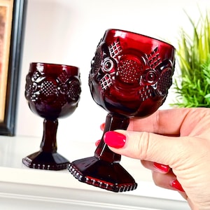 May include: Two deep red glass goblets featuring a raised geometric pattern on the bowl. Each goblet has a faceted stem and a square base with a beaded edge. The glass is translucent and dark red, showcasing a textured surface design throughout.