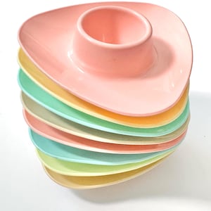 May include: A stack of eight triangular egg cups in pastel colors including pink, yellow, mint green, and cream. Each cup features a smooth, rounded rim and a central circular indentation designed to hold a single egg, arranged in a vertical column.