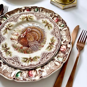 Peut inclure: Assiettes anciennes avec une illustration de dinde, entourée de motifs floraux et fruités marron, crème et vert. Comprend une fourchette et un couteau cuivrés. Les assiettes sont empilées, mettant en valeur le motif détaillé.