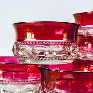May include: Several ruby red glass bowls with a clear, textured base are stacked. The bowls feature a decorative pattern of circles and a wide, red rim. The glass reflects light, creating a vibrant display of colour and texture.