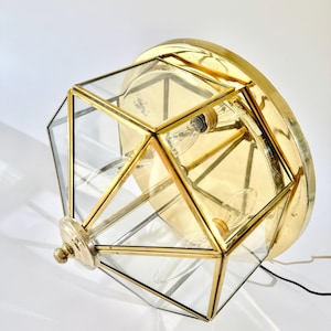 May include: A vintage brass and glass geometric ceiling light fixture. The fixture has a multi-faceted, clear glass shade with brass trim. The light has a round brass base and a small decorative finial. The light fixture is approximately 30 cm in diameter.