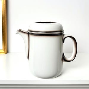 May include: A white ceramic teapot with a lid and a curved handle. The teapot has a dark brown trim around the edges and spout. The teapot is sitting on a white surface.