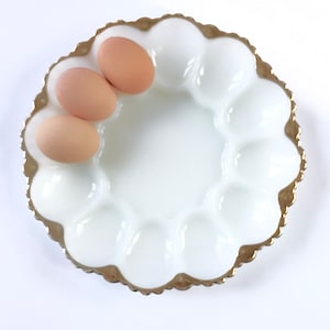 May include: A white, scalloped egg serving dish with a gold rim, holding three brown eggs. The dish has a central well surrounded by individual egg cups. The gold rim adds a touch of elegance to this kitchenware.