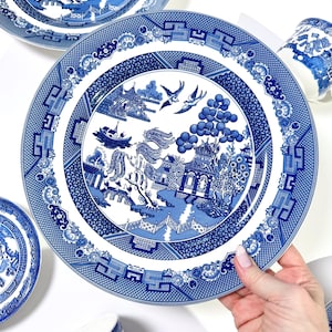 May include: A collection of blue and white Willow pattern plates and mugs. The plates display a detailed landscape scene with pagodas, bridges, and birds. The mugs have a matching design. Classic tableware.