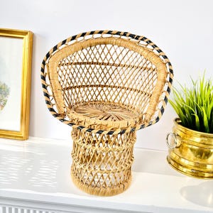 May include: A miniature peacock chair made from woven rattan. The chair has a rounded back and a cylindrical base. It is a natural tan colour with black detailing around the edge. A decorative accent piece.