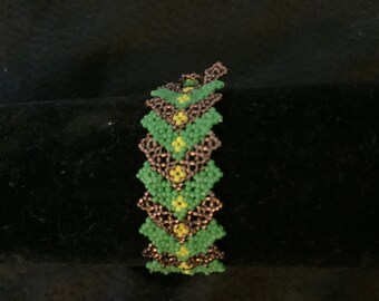 May include: A beaded bracelet with a zig-zag design. The bracelet is mainly green with yellow and bronze accents. The small beads are tightly woven, creating a textured surface. The bracelet is shown against a dark background.