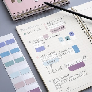 Könnte beinhalten: Ein Notizbuch mit einem rosa Einband und einer Spiralbindung. Das Notizbuch hat linierte Seiten und ist mit handschriftlichen Notizen in Chinesisch gefüllt. Es gibt auch mehrere farbige Haftnotizen mit leeren Feldern auf den Seiten. Die Haftnotizen sind in Blautönen, Grüntönen und Violett.