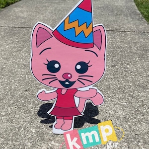 Puede incluir: Un gato rosa con un vestido rojo y un gorro de fiesta azul, amarillo y rojo. El gato tiene grandes ojos negros y una gran sonrisa. El gato est&aacute; de pie en una acera gris. El texto "kmp Party Crafts" es visible en la esquina inferior derecha de la imagen.