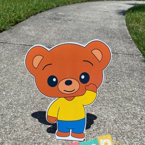 Puede incluir: Un personaje de dibujos animados de oso marr&oacute;n con una camisa amarilla y pantalones azules est&aacute; saludando. El personaje est&aacute; de pie en una acera gris.