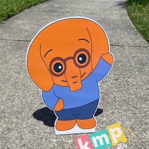 Puede incluir: Un recorte de personaje de dibujos animados de un elefante naranja que lleva gafas azules, una camisa azul y pantalones azules. El personaje est&aacute; saludando con su brazo derecho. El recorte est&aacute; en una acera gris con hierba verde en el fondo. El texto "kmp party crafts" es visible en la esquina inferior derecha de la imagen.