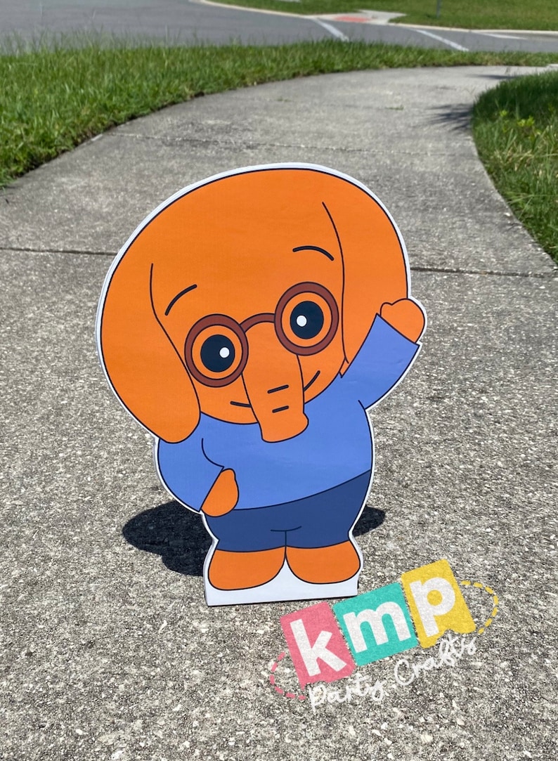 Puede incluir: Un recorte de personaje de dibujos animados de un elefante naranja que lleva gafas azules, una camisa azul y pantalones azules. El personaje est&aacute; saludando con su brazo derecho. El recorte est&aacute; en una acera gris con hierba verde en el fondo. El texto "kmp party crafts" es visible en la esquina inferior derecha de la imagen.