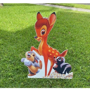 Peut inclure: Une découpe en carton de Bambi, le lapin Lapin et la belette Fleur du film Disney Bambi. Les personnages sont debout sur un fond blanc avec une bordure de marguerites blanches.