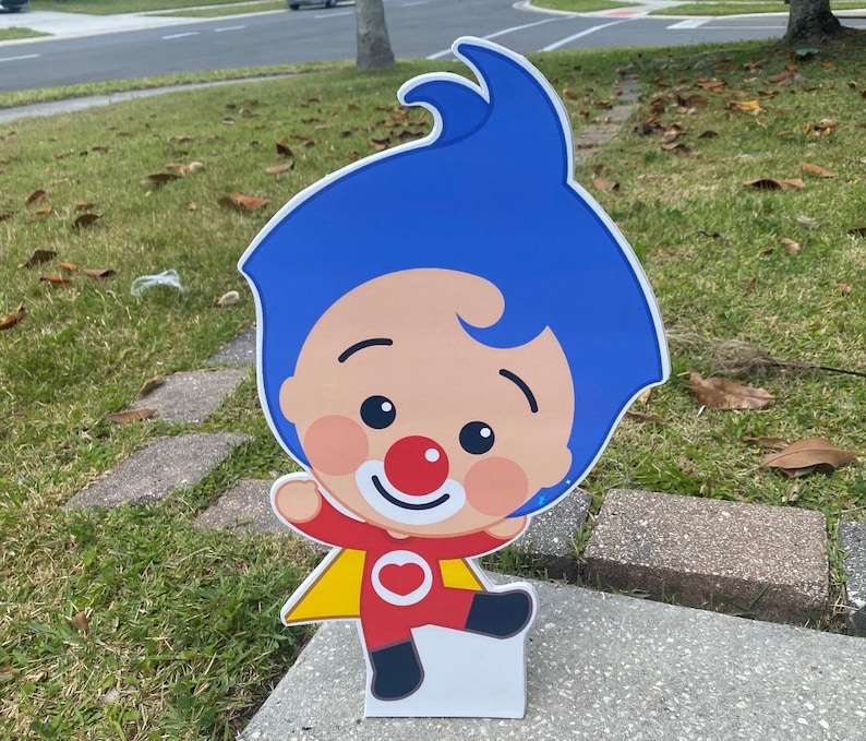 Puede incluir: Un recorte de cart&oacute;n de un payaso de dibujos animados con pelo azul, nariz roja y un traje rojo y amarillo. El payaso lleva una camisa roja con un coraz&oacute;n blanco y sostiene una capa amarilla.