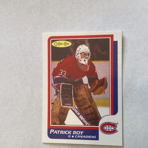 May include: A vintage O-Pee-Chee hockey card featuring Patrick Roy, a goalie for the Canadiens. The card has a red, white, and blue border. Roy is wearing a red jersey with the number 33 and a white helmet. The Canadiens logo is visible.