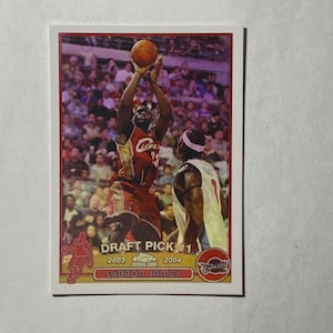 Puede incluir: Una tarjeta de baloncesto con LeBron James con el uniforme rojo y blanco de los Cleveland Cavaliers, a medio tiro. La tarjeta dice "DRAFT PICK #1 2003-2004 LeBron James Chrome Rookie Card" con el logo de Topps.