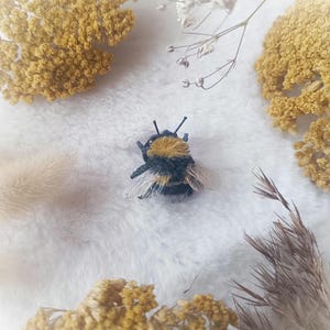 Peut inclure: Une figurine d'abeille faite à la main avec un corps jaune et noir duveteux, des ailes délicates et des antennes. L'abeille est entourée de fleurs jaunes séchées et de tissu blanc moelleux, créant une scène naturelle et fantaisiste.