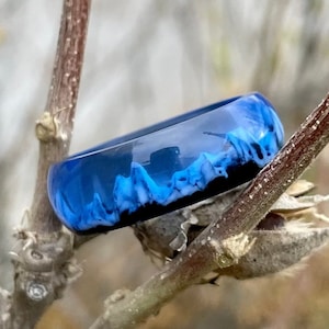 May include: A blue resin ring with a black base and a wave-like pattern of light blue and white. The ring is set against a blurred background of tree branches.