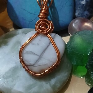 May include: A handcrafted pendant featuring a heart-shaped white stone with gray veining, wrapped in copper wire. The wirework includes a decorative spiral at the top. The pendant is displayed on a light green stone, with other colorful stones in the background.