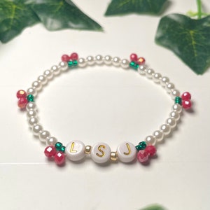 Op de afbeelding: Een witte parel armband met rode en groene kralen, die de naam "LSJ" spellen.