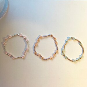 May include: Three beaded bracelets with flower-shaped charms. The bracelets are pink, beige, and light blue.