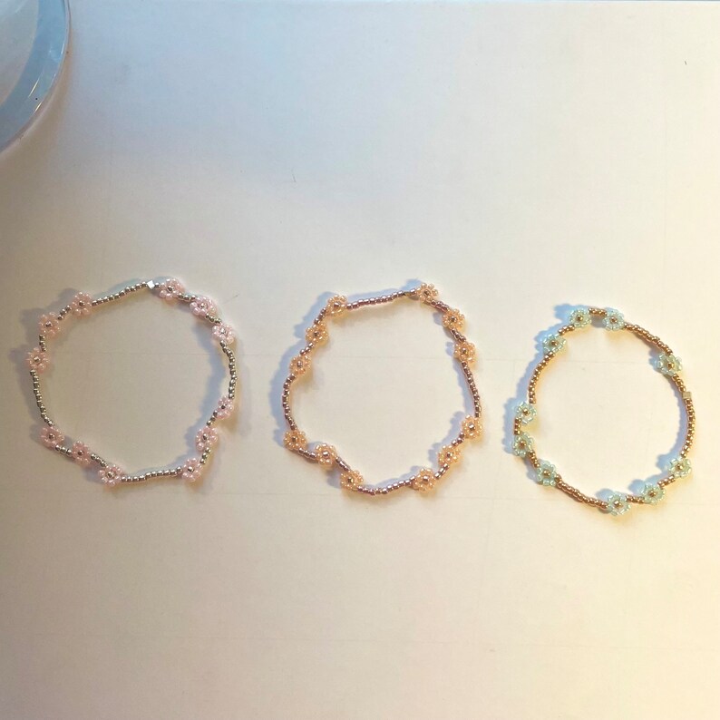 May include: Three beaded bracelets with flower-shaped charms. The bracelets are pink, beige, and light blue.