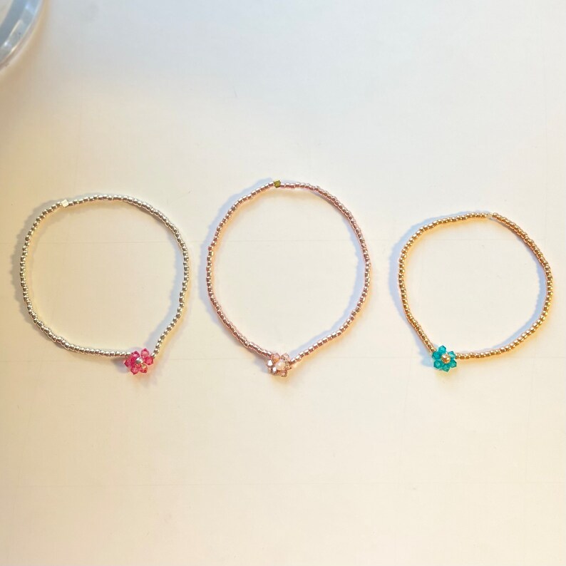 May include: Three delicate beaded bracelets with flower charms. The bracelets are silver, rose gold, and gold in colour. The flower charms are pink, light pink, and teal.
