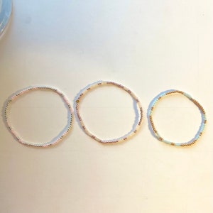 May include: Three beaded bracelets in different colours. The first bracelet is pink and white, the second is white and gold, and the third is light blue and gold.