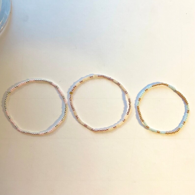 May include: Three beaded bracelets in different colours. The first bracelet is pink and white, the second is white and gold, and the third is light blue and gold.