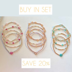 May include: Set of three beaded bracelets with flower charms. The bracelets are in pink, peach, and teal colours. The text "BUY IN SET SAVE 20%" is displayed above the bracelets.