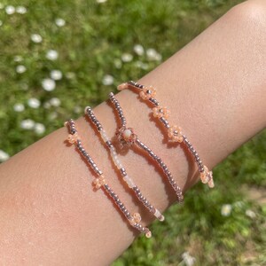 May include: Four delicate beaded bracelets with small flower accents in shades of pink and white. The bracelets are stacked on a person's wrist.