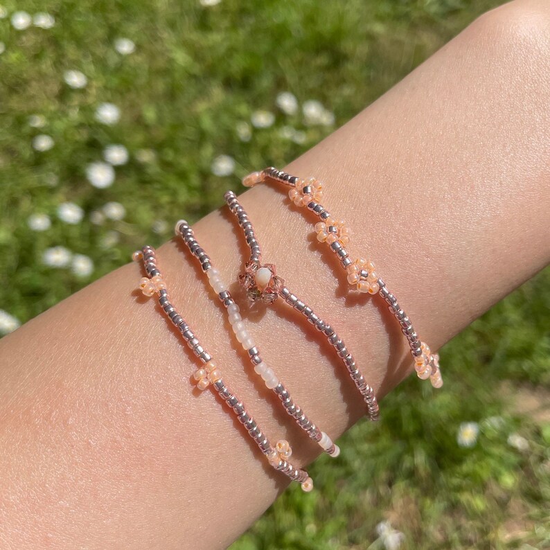 May include: Four delicate beaded bracelets with small flower accents in shades of pink and white. The bracelets are stacked on a person's wrist.