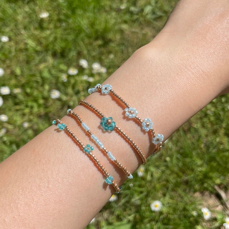May include: Four beaded bracelets with flower charms. The bracelets are made with light blue and gold beads. The flower charms are made with light blue and teal beads.