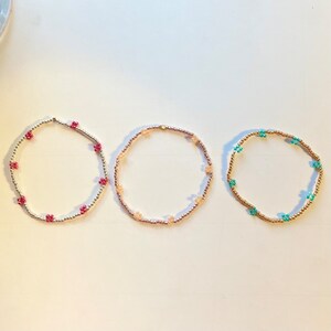 May include: Three beaded bracelets with different coloured flower charms. The bracelets are gold, pink and turquoise.