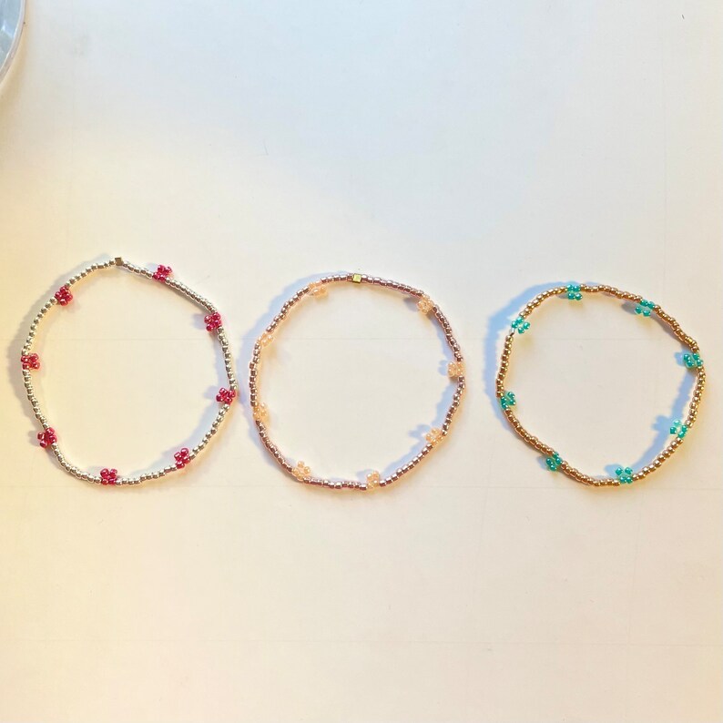 May include: Three beaded bracelets with different coloured flower charms. The bracelets are gold, pink and turquoise.
