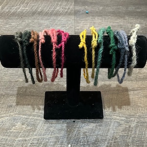 May include: A display of 10 colorful yarn bracelets on a black stand. The bracelets are made of different colors of yarn, including green, blue, yellow, pink, and brown.