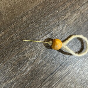 May include: A wooden bead with a natural wood grain pattern is strung on a thin, gold-colored wire. The wire is attached to a loop of natural twine.