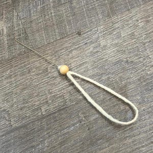 May include: A handmade needle threader with a wooden bead and a loop of white yarn. The needle threader has a thin, silver wire for threading needles. The wooden bead is light brown.