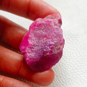 May include: A rough, uncut ruby gemstone, held in a hand. The ruby is a vibrant shade of pinkish-red with a textured surface. The gemstone's irregular shape and natural appearance are prominent.