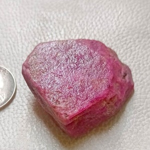 May include: A rough, uncut ruby gemstone with a deep pinkish-red hue. The ruby is next to a U.S. quarter for size comparison. The gemstone has a textured surface and irregular shape, showcasing its natural form. Ideal for jewelry making or gemstone collectors.